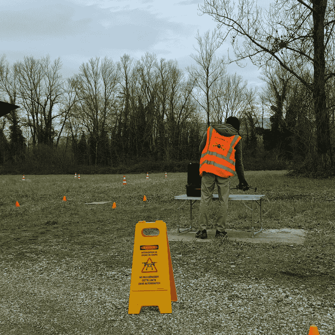 Une formation drone professionnel en extérieur sur un terrain de vol, on aperçoit le formateur et une zone balisée avec un panneau jaune d'avertissement.